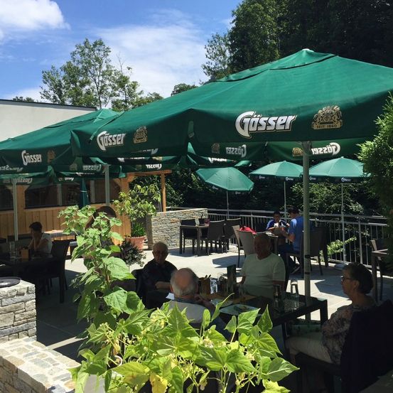 Restaurant im Freien mit großen grünen Sonnenschirmen. Leute sitzen unter den Schirmen und genießen Getränke. Pflanzen und Bäume sind in der Umgebung.