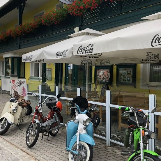 Eine Straßenszene mit mehreren Vintage-Mopeds, die vor einem Café mit Coca-Cola-Schirmen geparkt sind, roten Blumen auf dem Balkon und einem Eiscreme-Schild.
