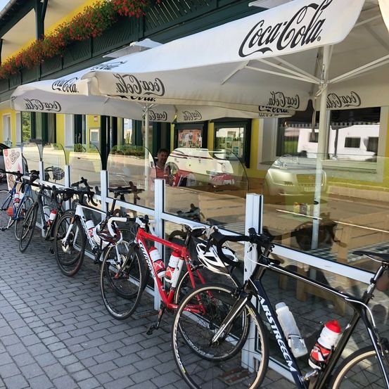 Fahrräder sind vor einem Café mit Coca-Cola-Schirmumbrellas geparkt. Eine Person spiegelt sich im Fenster wider, mit einem geparkten Auto im Hintergrund.