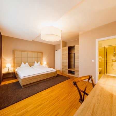 Ein elegant gestaltetes Schlafzimmer mit Holzboden, einem Doppelbett und einem hölzernen Kopfteil. Das Zimmer verfügt über zwei Nachttische mit Lampen und einen hölzernen Stuhl. Das angrenzende Badezimmer hat eine Glasdusche und Toilette.