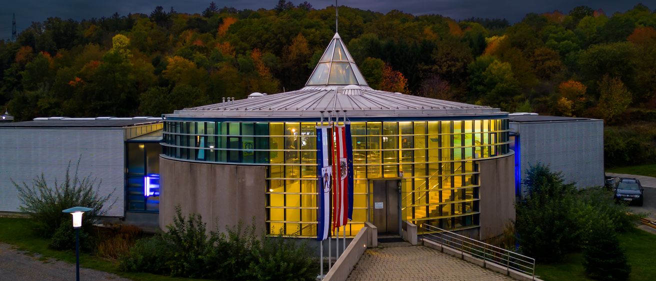 Ein modernes Gebäude mit einem Glaskuppeldach, das nachts beleuchtet ist. Flaggen sind auf Stangen davor angebracht. Das umliegende Gebiet ist mit grünen Bäumen gefüllt.