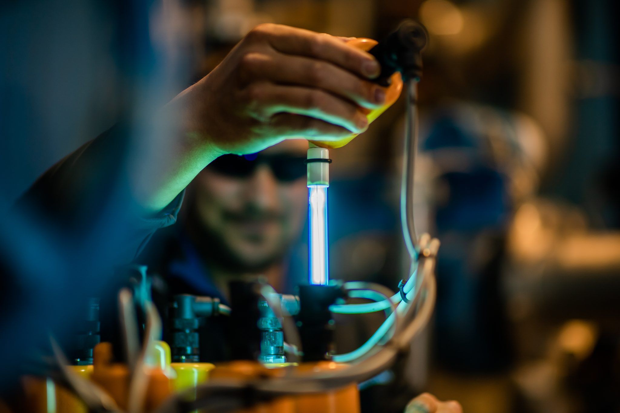 Eine Person hält eine leuchtend blaue Lampe, vielleicht ein wissenschaftliches Experiment. Die Lampe ist mit einem orangefarbenen Objekt mit Kabeln verbunden.