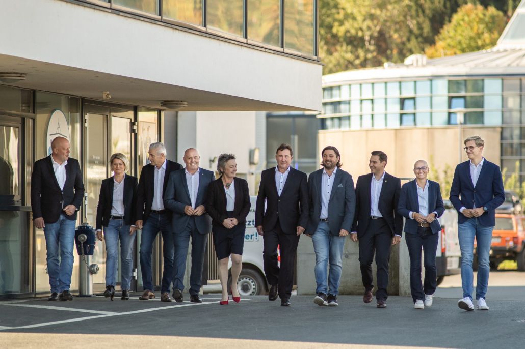 Eine Gruppe von Personen in formeller Kleidung geht vor einem modernen Gebäude. Im Hintergrund befinden sich Bäume und ein weiteres Gebäude.