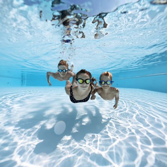 Drei Kinder schwimmen mit Schwimmbrillen und Schwimmkappen unter Wasser in einem Pool. Eine Frau lächelt und schaut zur Kamera.
