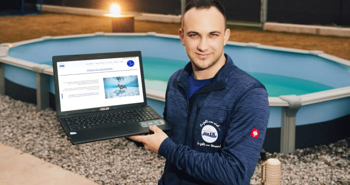 Ein Mann in einem blauen Jacke hält einen Laptop vor einem oberirdischen Pool. Der Laptop-Bildschirm zeigt eine Website mit Text und einem Bild.