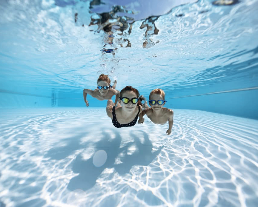 Drei Kinder mit Schwimmbrillen schwimmen in einem Freibad, wobei ein blauer Himmel über dem Wasser sichtbar ist.