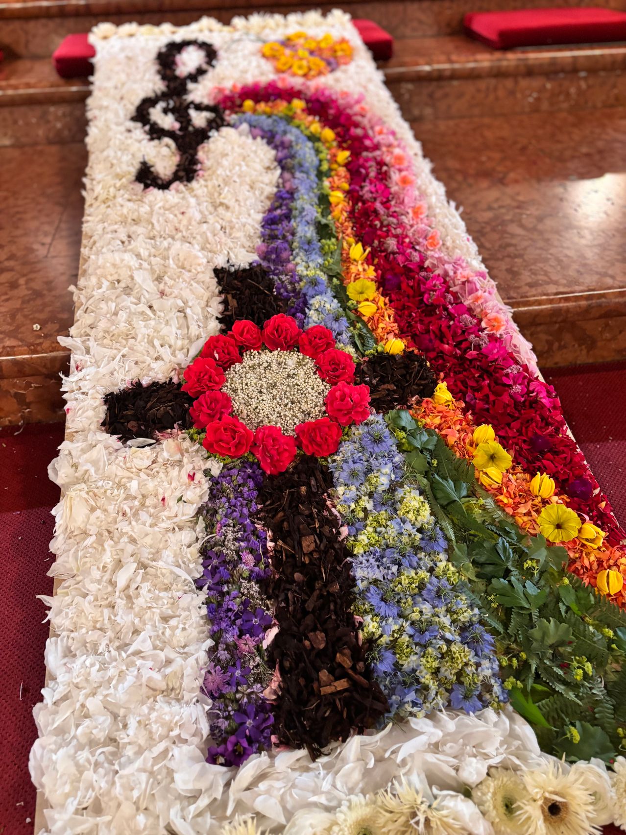 A vibrant floral carpet displays a cross with red roses, surrounded by a rainbow of colorful flowers. The carpet is on a wooden surface.