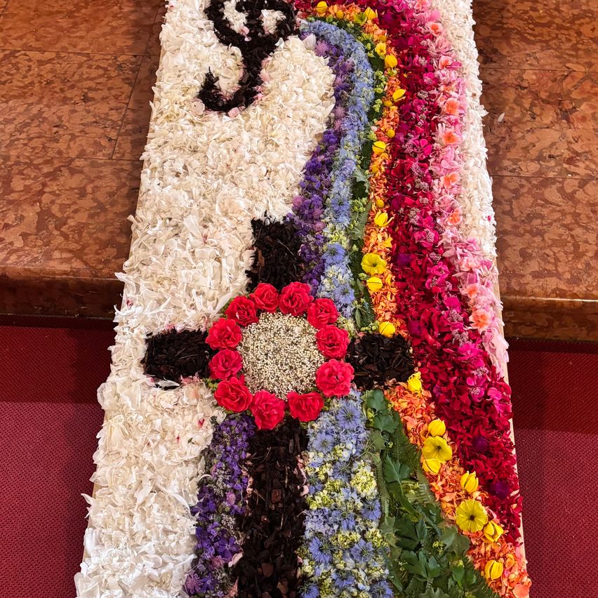 A colorful floral arrangement forms a heart shape on a white background, surrounded by various flowers. The floor beneath is tiled, with a red carpet on top.