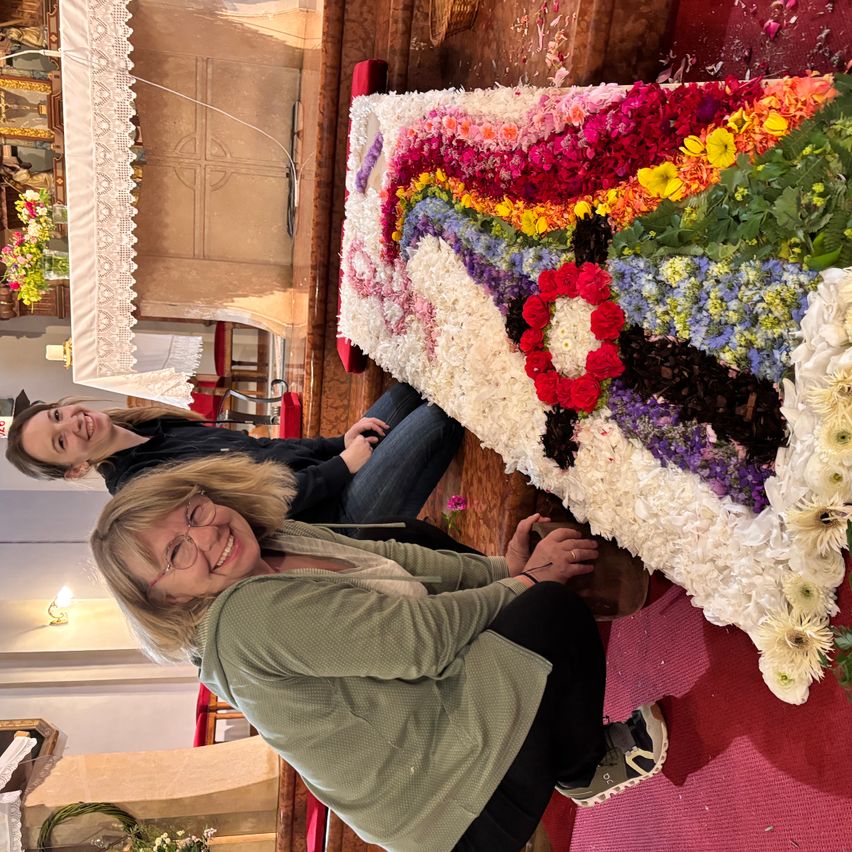 Two women smiling and sitting beside a colorful floral arrangement on a wooden surface. The arrangement includes roses and other flowers. Behind them, a wooden door and a white cloth.