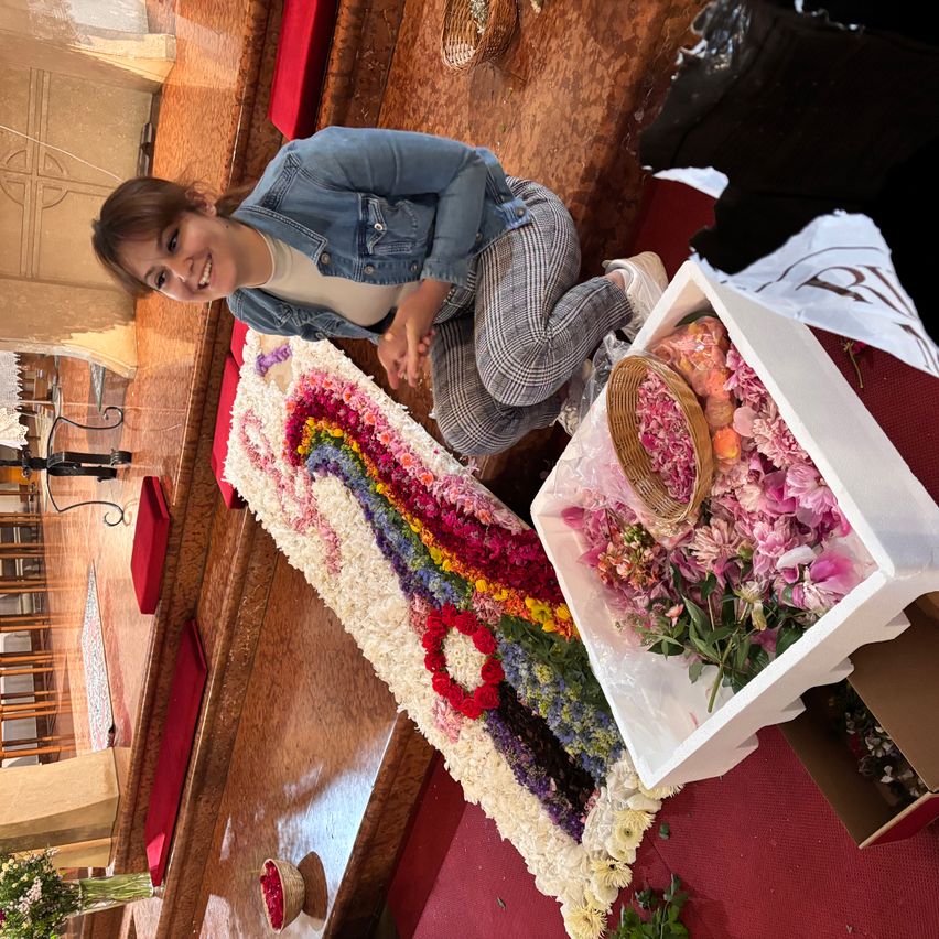 A woman in a denim jacket sits beside a large floral arrangement on a church floor, smiling at the camera. Nearby, a box of flowers is open on a red carpet.