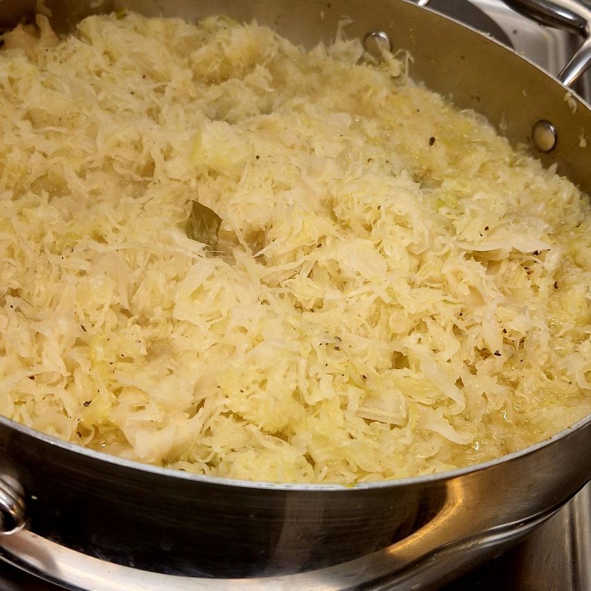 Ein Nahaufnahme von Sauerkraut, das in einer Metallpfanne auf einem Herd gekocht wird, wobei einige Kohlstücke sichtbar sind.