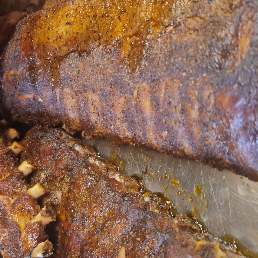 Nahaufnahme von geräucherten Rippen mit einer glänzenden, karamellisierten Außenseite, überzogen mit Soße und tropfend vor Säften. Das Fleisch ist zart und saftig, mit sichtbaren Knochen und einem verkohlten, rauchigen Geschmack.