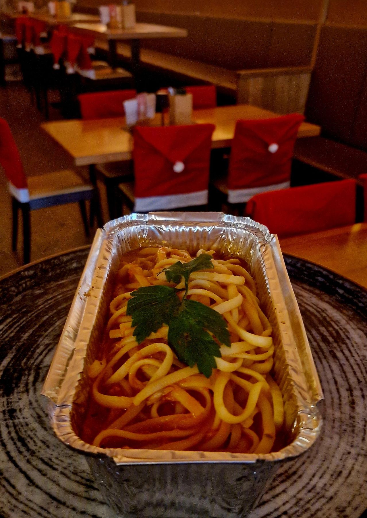 Eine Nahaufnahme einer Folie abgedeckten Pasta-Schale mit roter Sauce, garniert mit Petersilie, auf einem schwarzen Teller. Dahinter sind Stühle und Tische mit roten Sitzbezügen zu sehen.