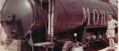 Ein alter schwarzer Tankwagen mit dem Wort 'Mobil' darauf. Ein Mann in Shorts steht daneben, trägt ein weißes Hemd und lächelt.