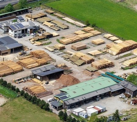 Luftaufnahme eines Sägewerks mit mehreren Holzstapel, Gebäuden und einer Straße. Das Gebiet ist von Grün umgeben.
