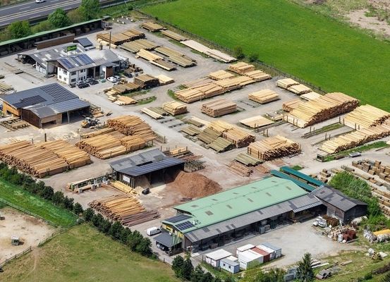 Luftaufnahme eines Sägewerks mit mehreren Holzstapel, Gebäuden und einer Straße. Das Gebiet ist von Grün umgeben.