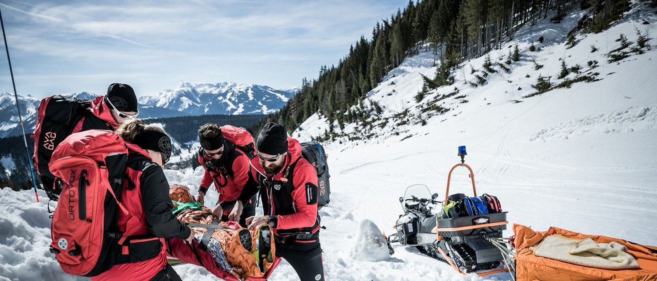 Ein Team von Rettern in roter Ausrüstung hilft einer Person auf einer Trage in einem verschneiten Berg. Ein Schneemobil mit Ausrüstung steht in der Nähe.