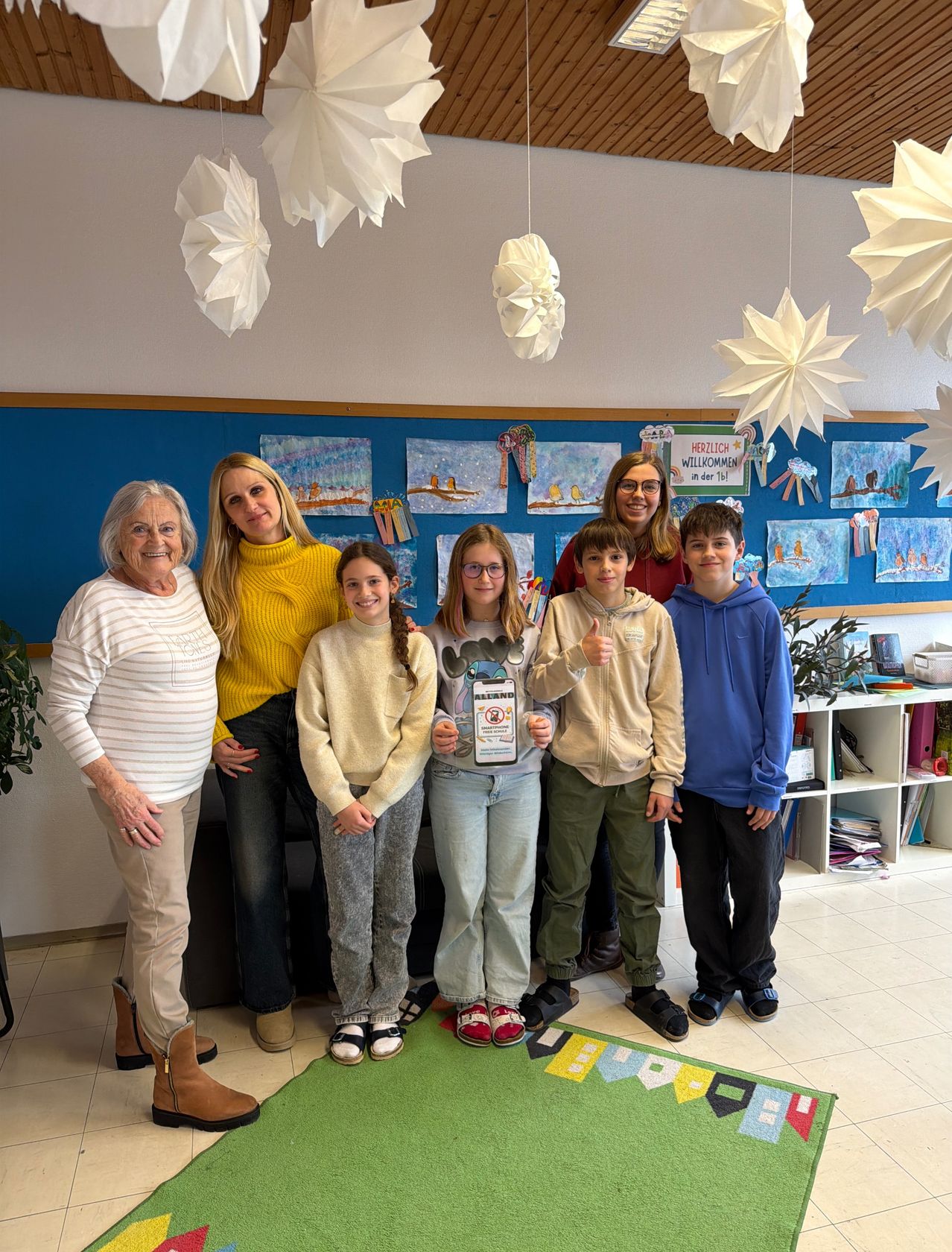 Eine Gruppe von Menschen steht in einem Klassenzimmer mit Dekorationen. Zwei Mädchen und ein Junge halten ein Zertifikat. Eine Frau trägt eine Brille und eine andere einen Ring.