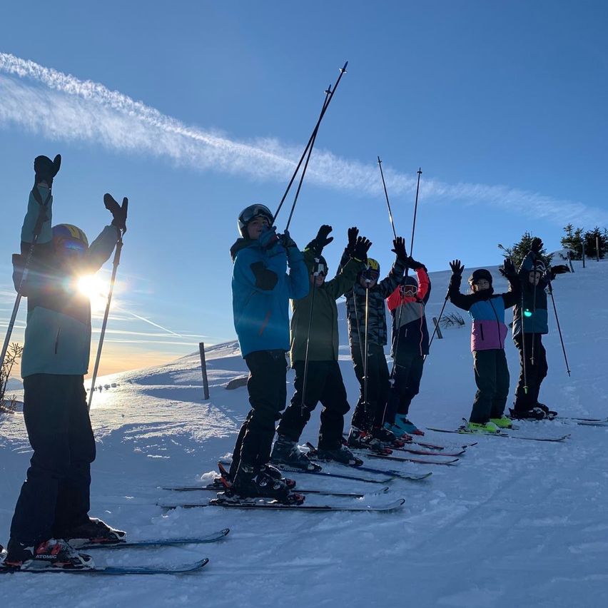 Eine Gruppe von Skifahrern posiert auf einem verschneiten Berg, hält ihre Skistöcke und trägt Helme. Die Sonne geht im Hintergrund unter und wirft ein warmes Licht.