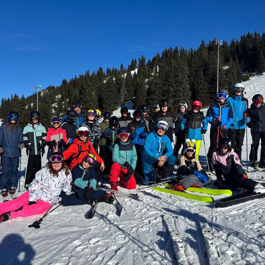 Eine Gruppe von Skifahrern und Snowboardern posiert für ein Foto auf einem verschneiten Berg. Einige stehen, andere sitzen und alle tragen Winterkleidung und Helme.