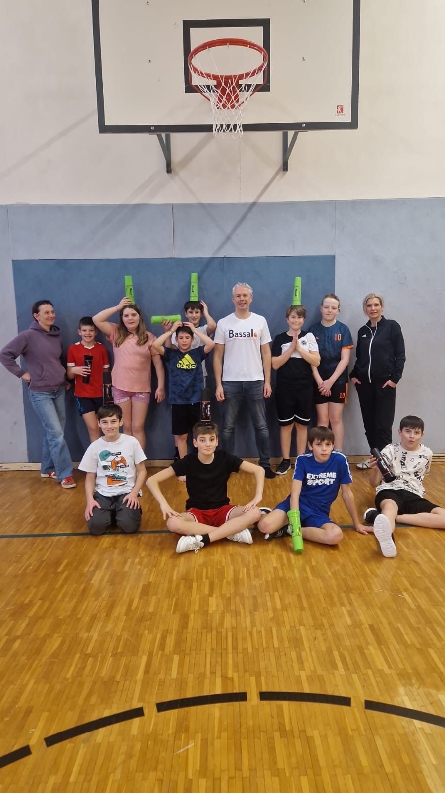 Eine Gruppe von Kindern und Erwachsenen posiert für ein Foto in einer Turnhalle und hält grüne Stöcke. Der Mann in der Mitte trägt ein weißes Shirt mit dem Wort Bassal darauf.