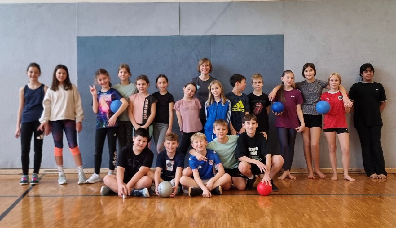 Eine Gruppe junger Leute mit verschiedenfarbigen Shirts posiert für ein Foto in einer Turnhalle mit Holzböden.