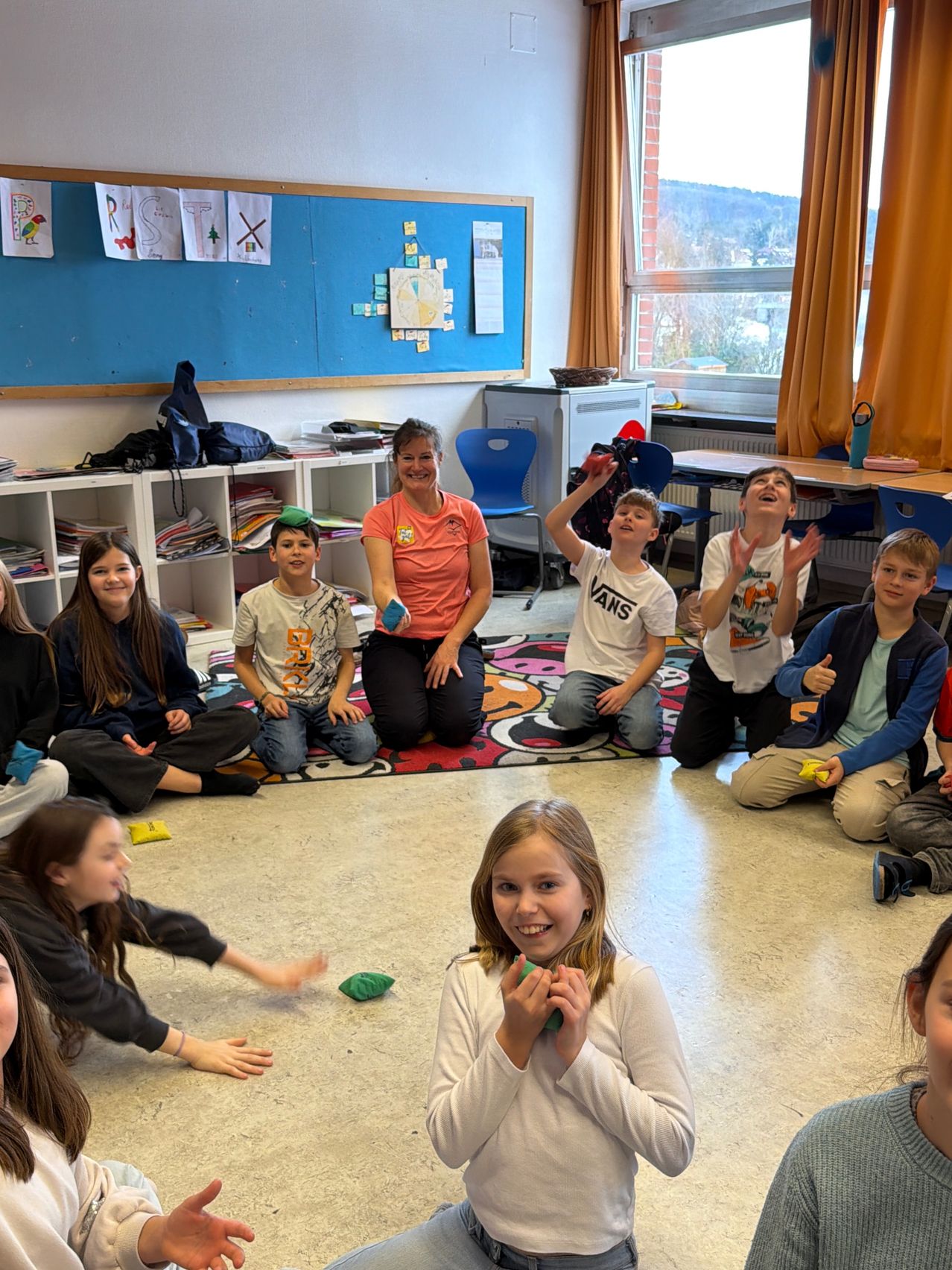 Ein Klassenzimmer mit Kindern auf dem Boden sitzend, eine Frau kniend und lächelnd, ein blauer Bulletin-Board mit Papieren, ein Heizkörper und Fenster mit Vorhängen, die einen Blick nach draußen zeigen.