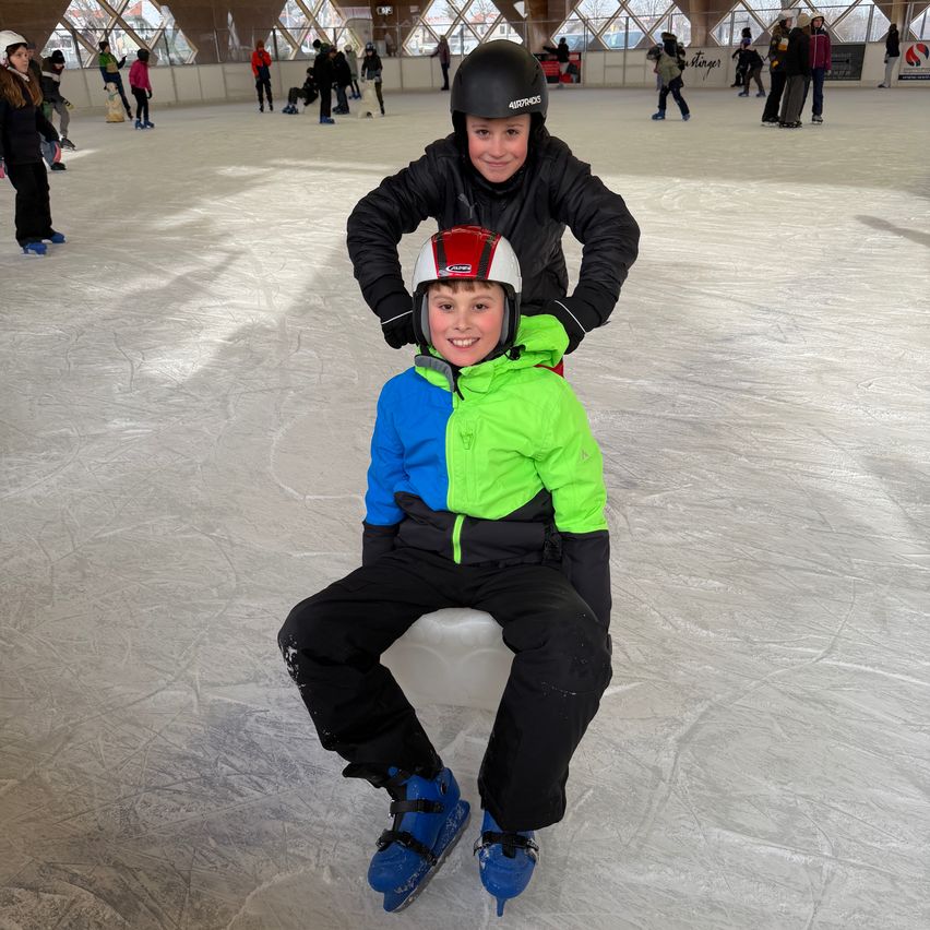 Zwei junge Jungen sind auf einer Eisbahn. Ein Junge sitzt auf einer Bank, während der andere hinter ihm steht, beide tragen Winterkleidung und Helme.