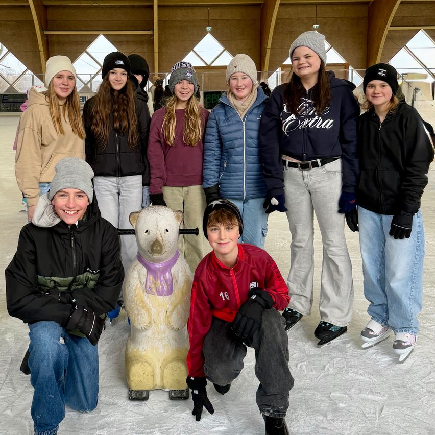 Eine Gruppe lächelnder Teenager auf einer Eisbahn, einige in Winterkleidung, posieren mit einer großen Eisbärenstatue. Der Hintergrund zeigt eine moderne Eisbahn mit Holzbalken und Glaswänden.