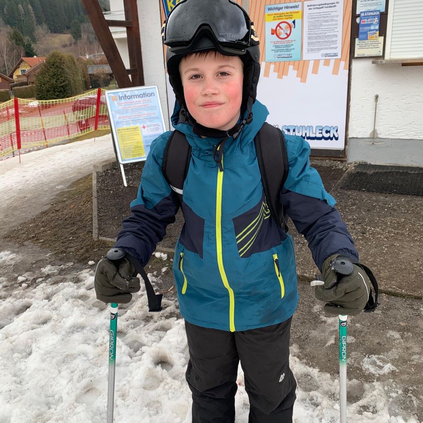 Ein junger Junge steht im Schnee, trägt eine blaue Jacke, eine Brille und Skistöcke. Hinter ihm steht ein Schild mit Informationen und ein Gebäude.