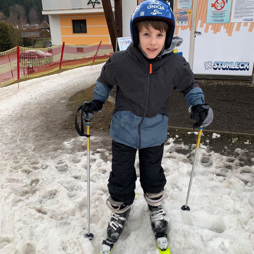 Ein junger Junge steht im Schnee und skifahrer, trägt einen blauen Helm, Skiausrüstung und lächelt. Hinter ihm steht ein Gebäude mit einem Schild, das 'STUHLECK' und einen Zaun zeigt.