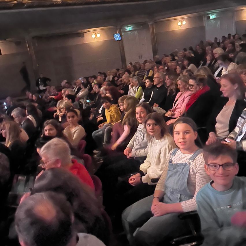 Ein großes Publikum sitzt in einem Theater, viele tragen Brillen und Freizeitkleidung. Ein Mädchen in einer Jeans-Latzhose lächelt. Ein Junge mit Brille sitzt neben ihr. Eine kleine Fernsehbildschirm ist an der Wand montiert.