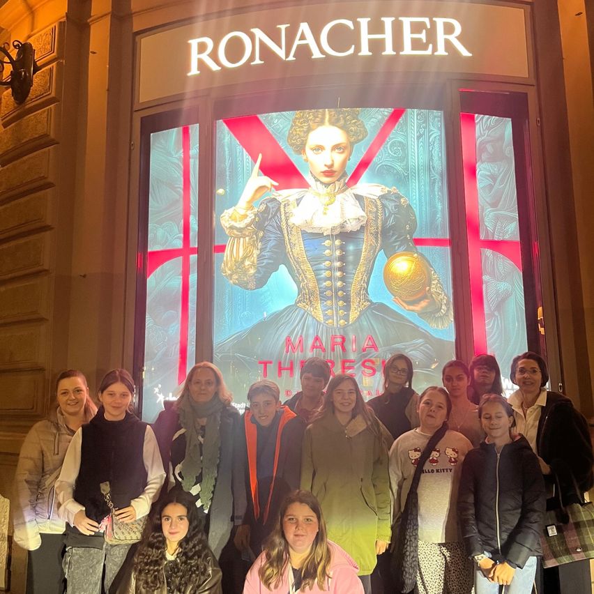 Eine Gruppe von Menschen steht vor einem Geschäft namens Ronacher, wobei ein großes Bild einer Frau in historischer Kleidung auf der Glastür zu sehen ist.