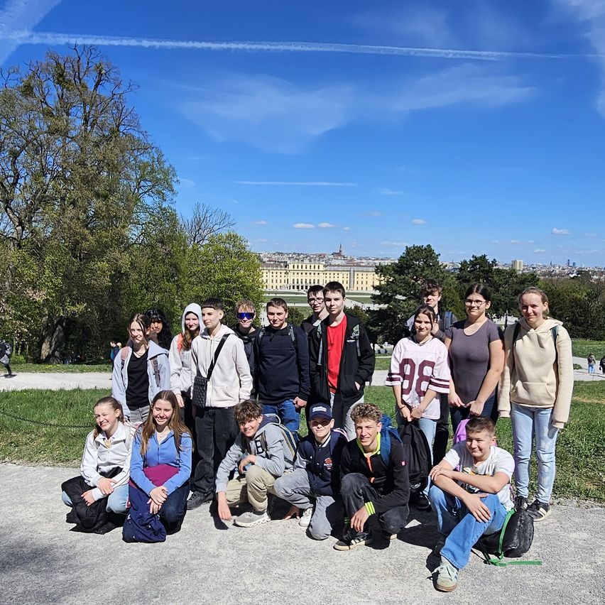 A group of young people, including both boys and girls, are posing for a photo in front of a historic building. They are standing on a pathway with trees and grass in the background.