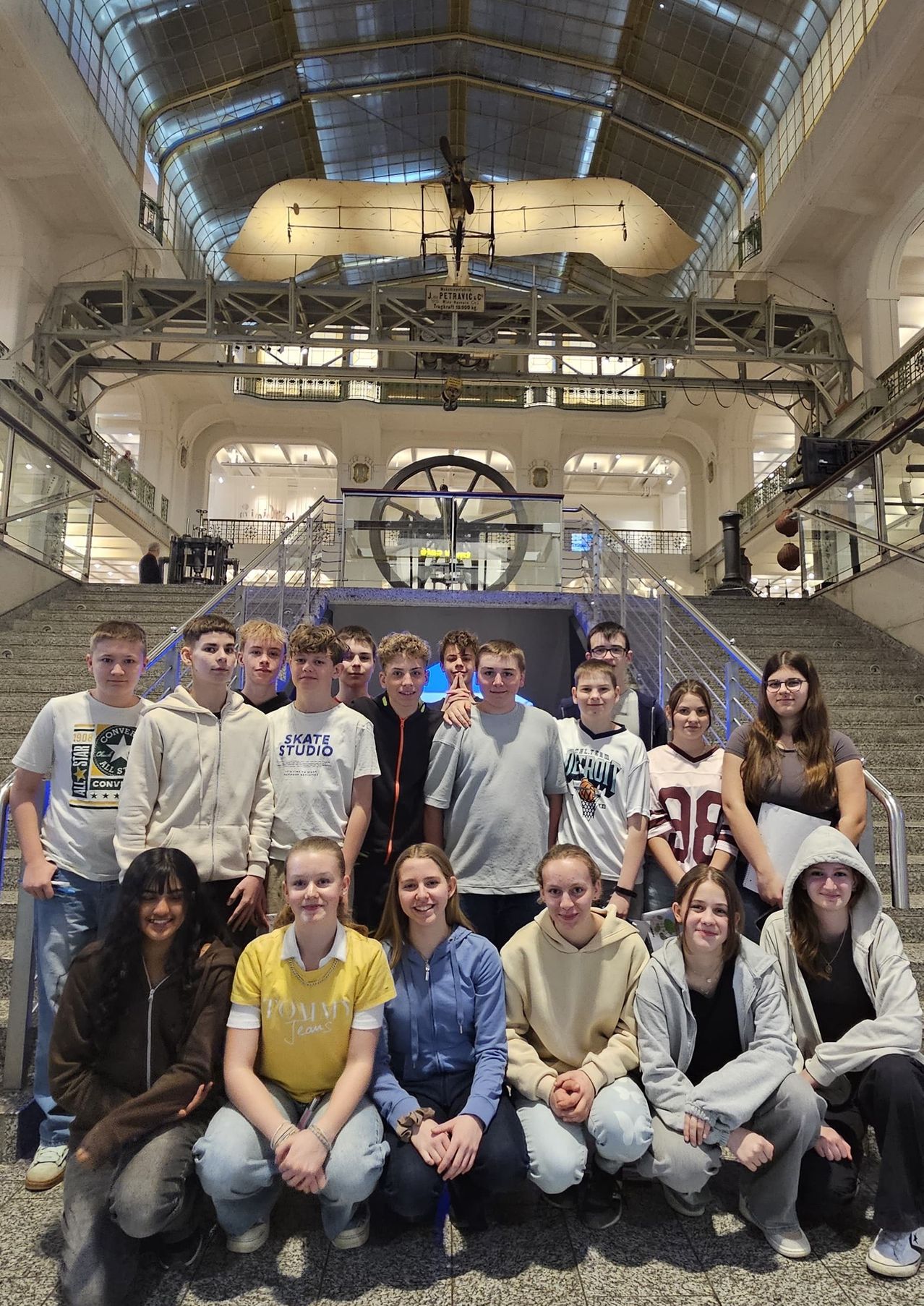 A group of teenagers, both boys and girls, are posing for a photo on the stairs inside an indoor facility. They are all smiling, and some of them are wearing glasses. Behind them, there is a large metal structure and a glass railing.