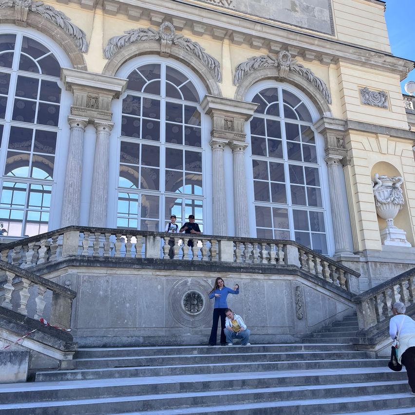 People pose in front of a grand, classical building with arched windows and a decorative balcony. Two individuals stand on the balcony while others crouch on the steps.
