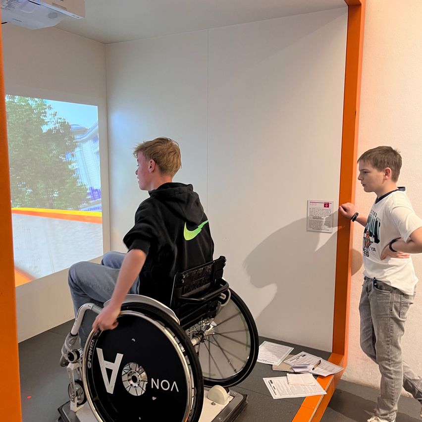 Two boys in a room with a large projection. One boy is in a wheelchair, and the other is standing next to him. Papers are scattered around.