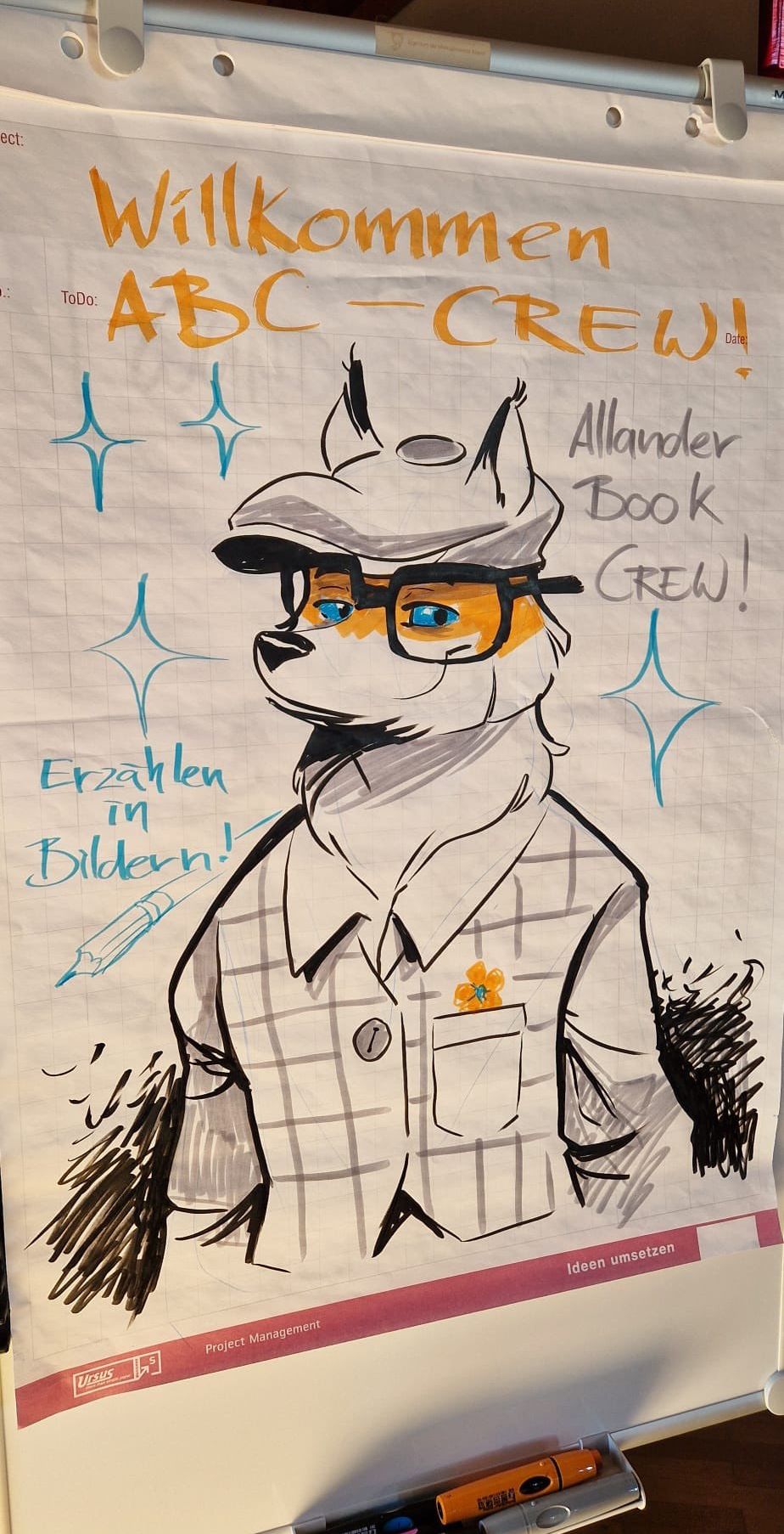 A drawing of a fox wearing a hat and glasses, with a checkered shirt and a flower on the pocket. The words 'Erzahlen in Bildern!' and 'Allander Book Crew!' are written on the paper.