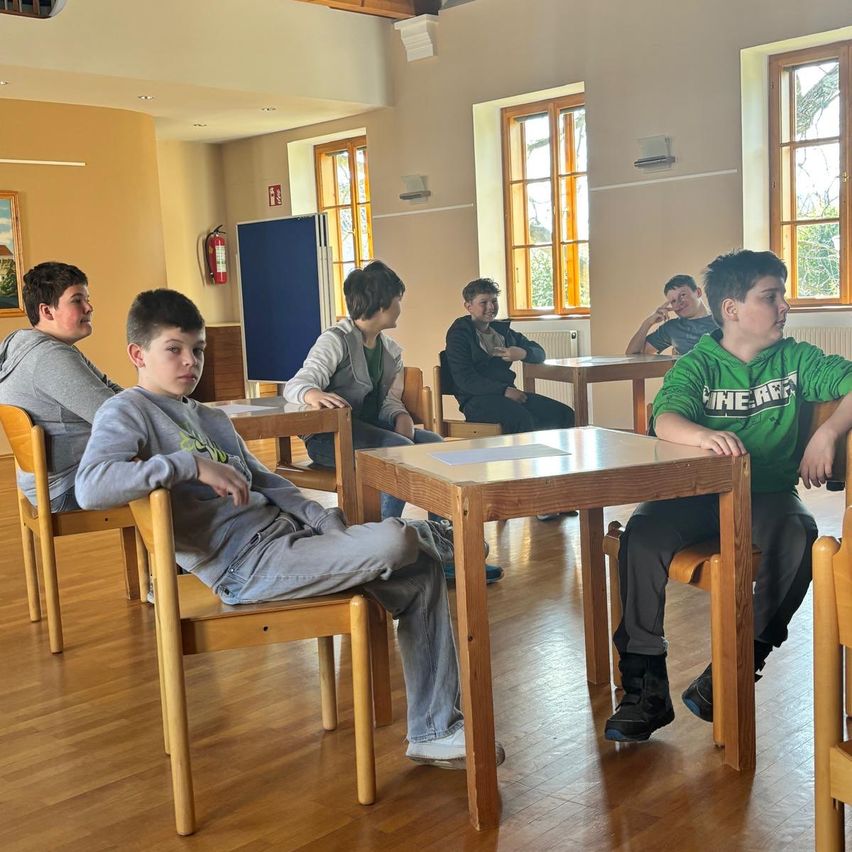 Six boys sit at tables in a classroom, some engaged in conversation, others with thoughtful expressions. The room has wooden floors, yellow walls, and windows with white frames.