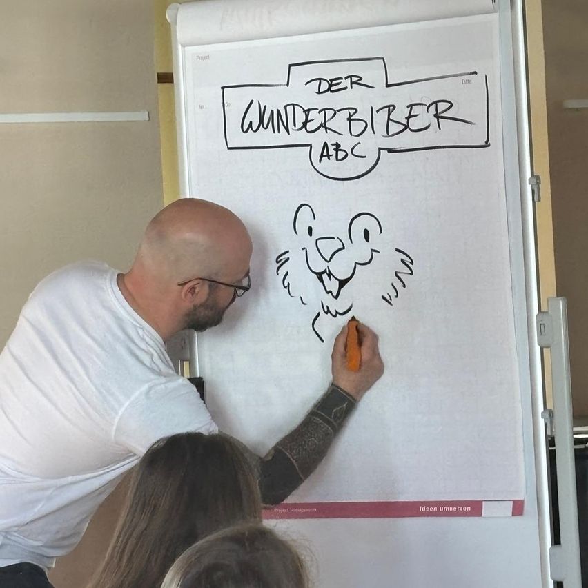 A man is drawing a cartoon of a bear on a whiteboard labeled 'Der Wunderbiber ABC'. Two women are observing the drawing.