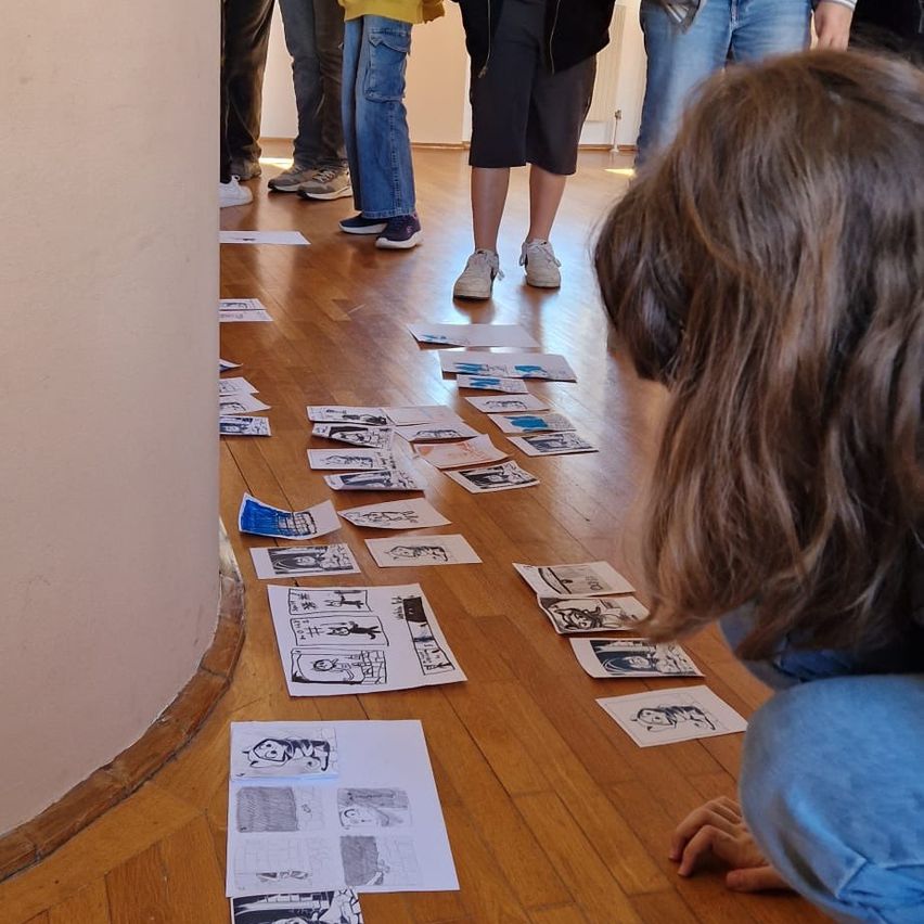 A group of people are gathered around in a room, looking at comic strips on the floor. A girl is kneeling and examining the drawings closely.