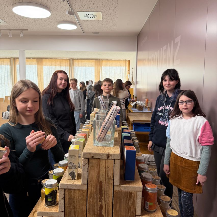 A group of young girls stand at a counter with various jars and boxes. The wall behind them has the word 'Brechenz' written on it. They are smiling and looking at the items.