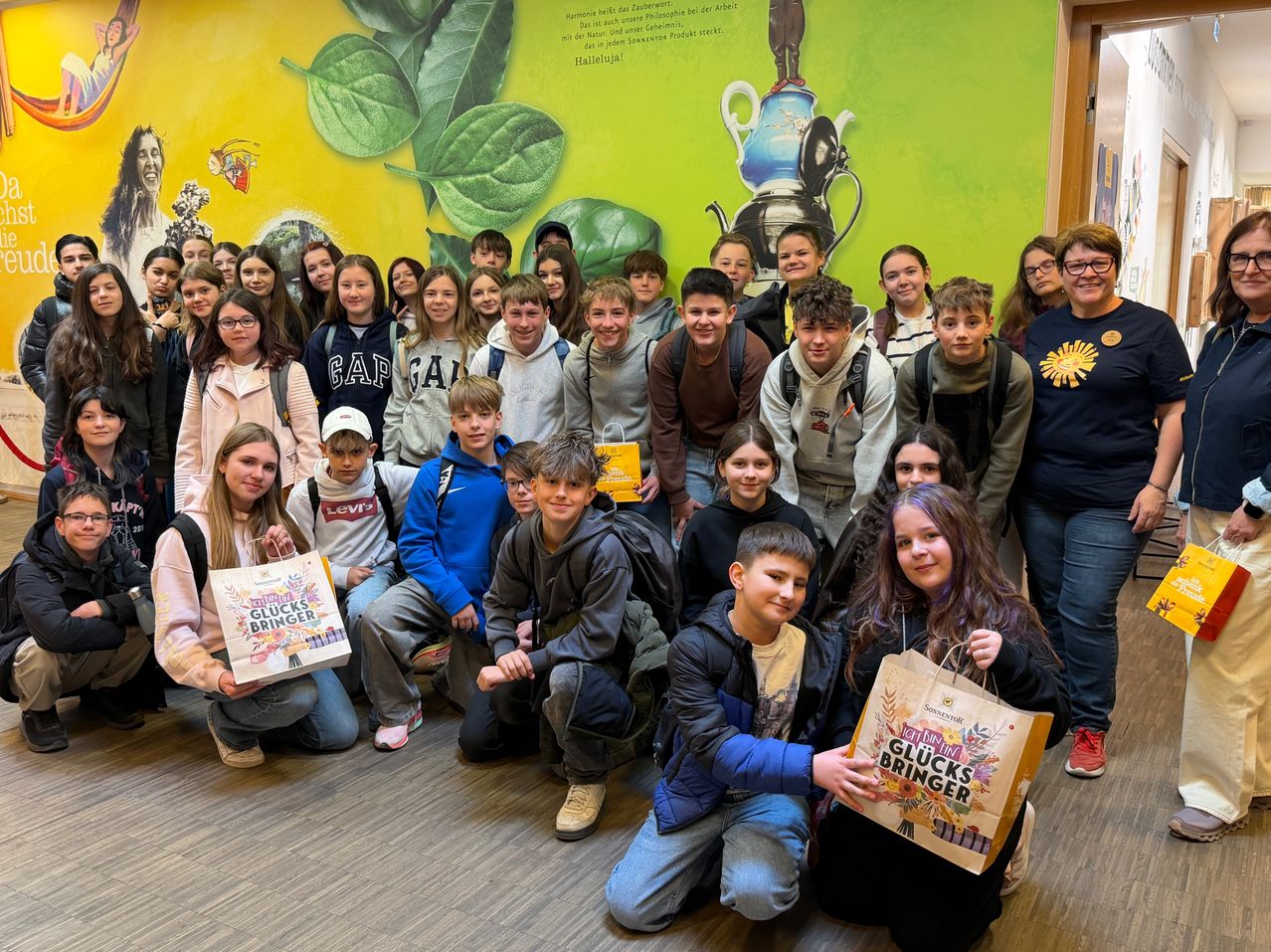 Group photo of students holding bags. A green wall with leaves and a teapot is in the background.