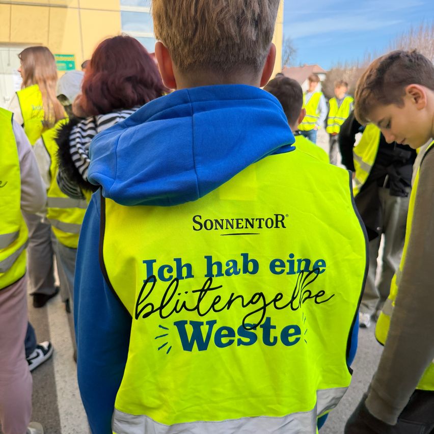 A boy in a blue hoodie with a yellow vest that says 'Ich hab eine Blütengelbe Weste' stands among a group of people in reflective vests.
