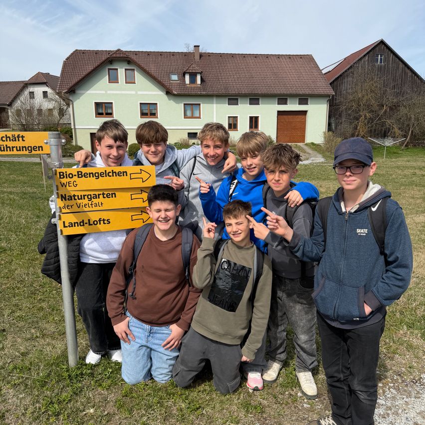A group of young boys pose for a picture in front of a house with a sign indicating directions to Bio-Bengelchen, Naturgarten der Vielfalt, and Land-Lofts.
