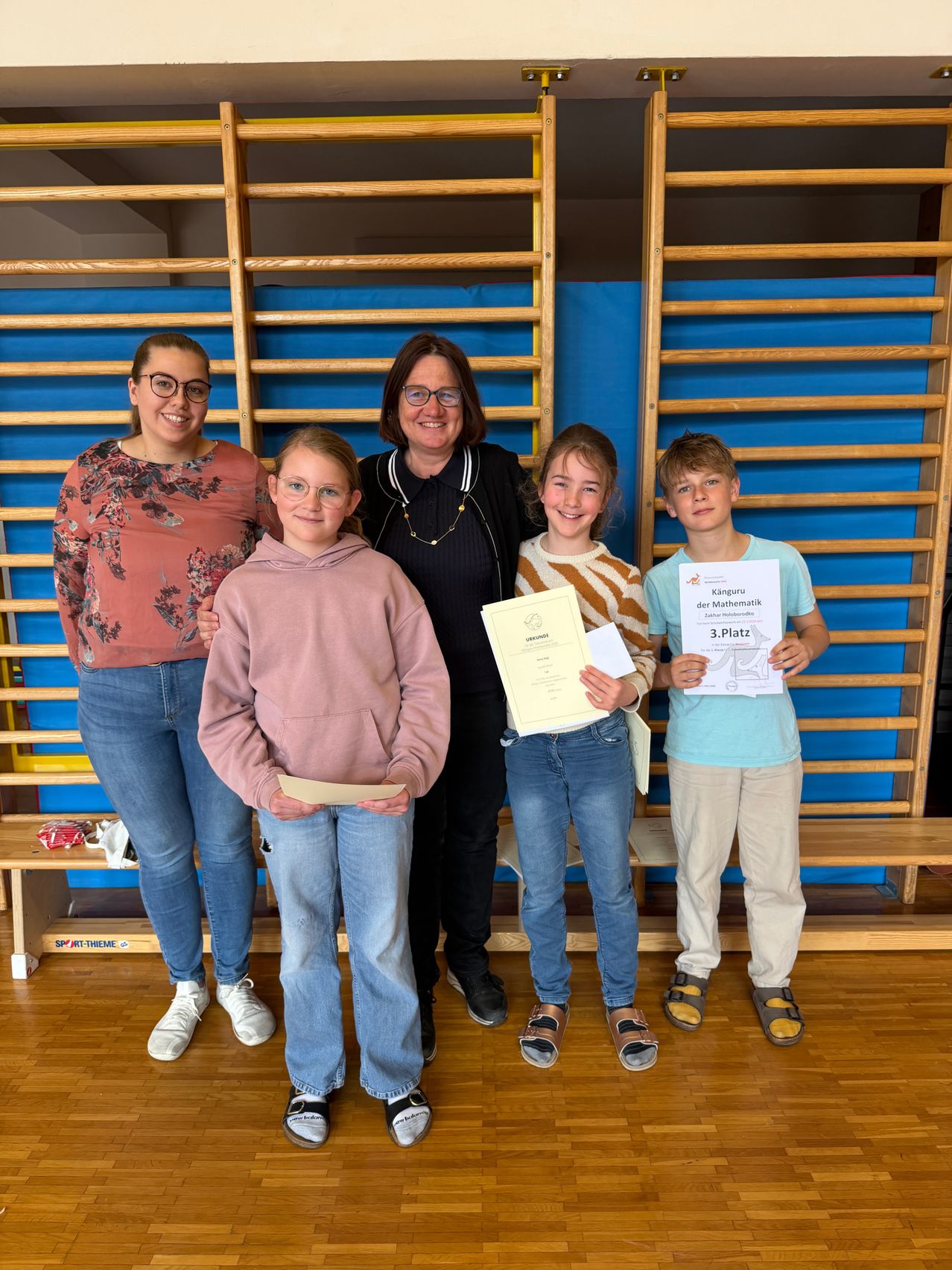 Five people stand in a room. Three young people hold certificates. One woman and one young person smile. They stand in front of wooden structures.