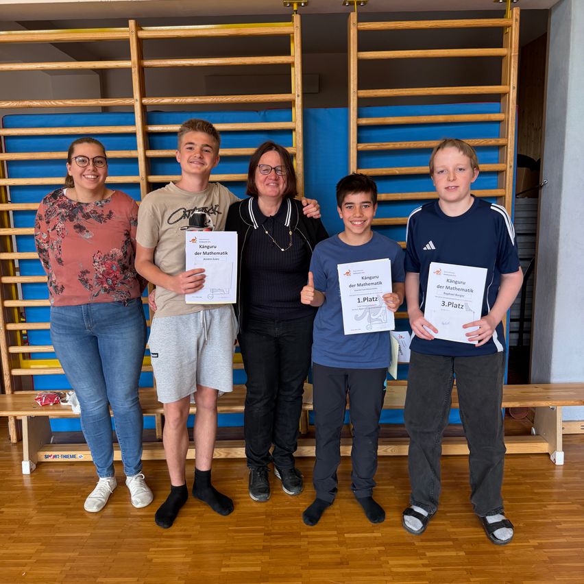 Five people are standing in a room, holding certificates. A woman is holding a boy on her shoulder. The boy in the middle is wearing a shirt with text and numbers on it. The woman on the right is wearing glasses and a necklace. Behind them is a blue wall.