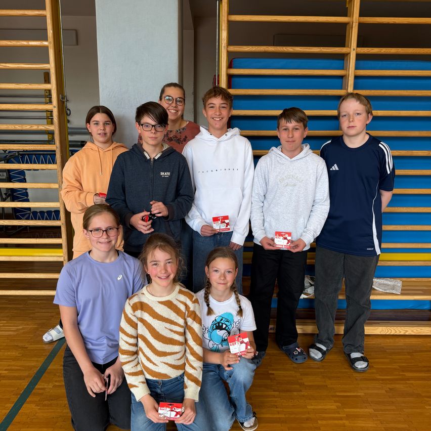 A group of young individuals, some standing and others crouching, pose for a photograph in a gymnasium. They hold small, red items and wear casual clothing. Behind them, wooden ladders and blue foam mats are visible.