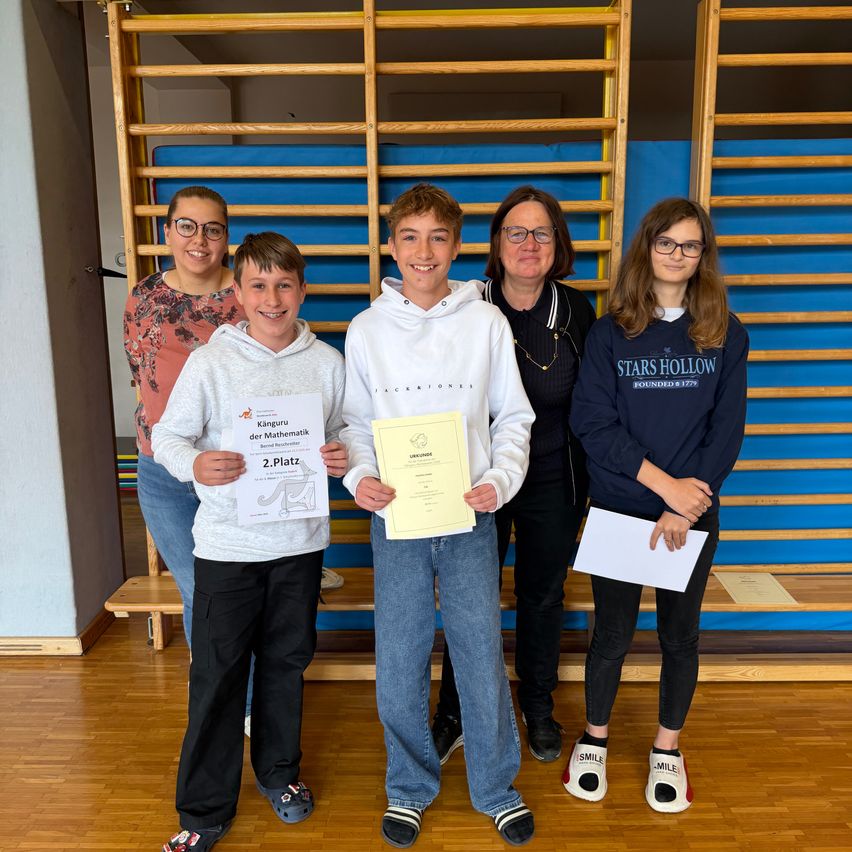 Five individuals stand in a room with wooden floors and blue walls. Three are holding certificates, and two are wearing glasses. The boy in the middle wears a white hoodie.