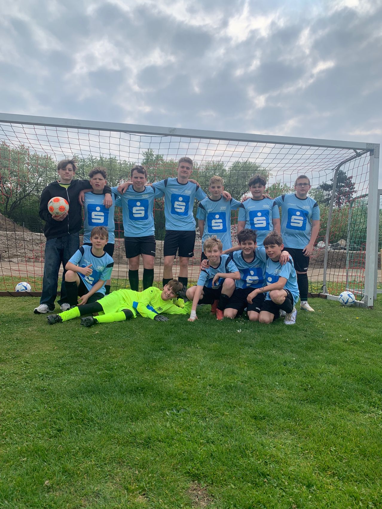 A youth soccer team poses for a photo in front of a goal, with players in blue jerseys and one holding a soccer ball.